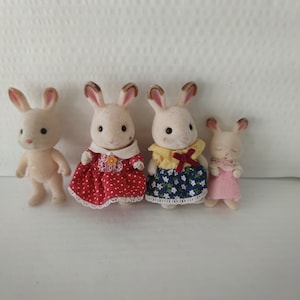 May include: Four white rabbit figurines with different colored dresses. The rabbit on the left is wearing a plain white dress, the next rabbit is wearing a red dress with white polka dots, the third rabbit is wearing a blue dress with white flowers, and the last rabbit is wearing a pink dress.