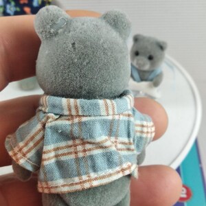 May include: A grey teddy bear figure wearing a blue and brown plaid shirt. The bear has small, round ears and a button nose. Other similar bear figures are in the background.