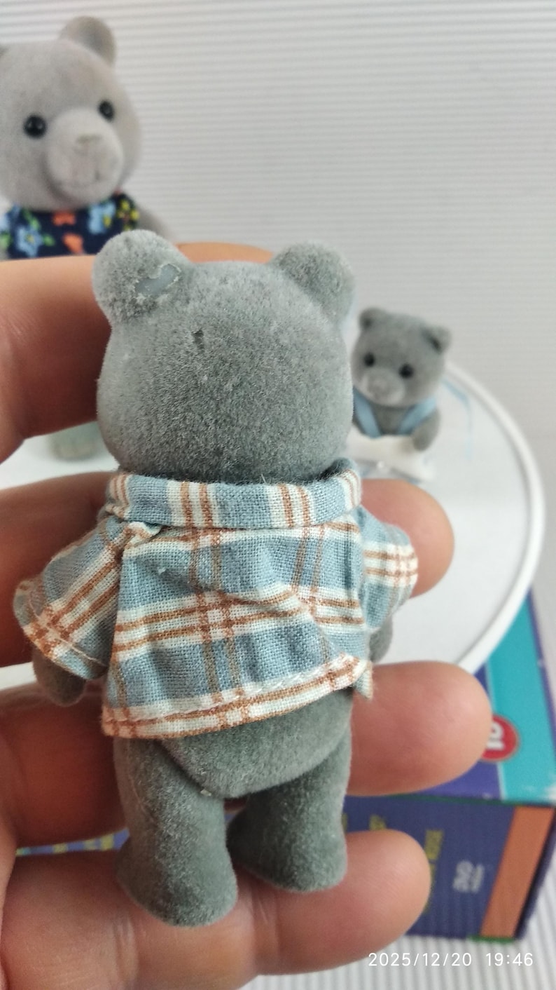May include: A grey teddy bear figure wearing a blue and brown plaid shirt. The bear has small, round ears and a button nose. Other similar bear figures are in the background.
