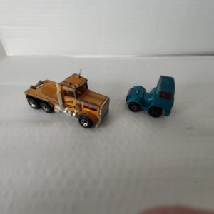 May include: Two toy semi-trucks. One is yellow and orange with silver accents and black wheels. The other is a small blue truck. Both are on a white surface.