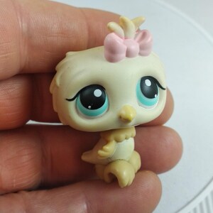 May include: A small, collectible bird figurine with a cream-colored body and large, expressive eyes. It has a pink bow on its head and a yellow beak. The toy is held in a hand, suggesting its small size and collectible nature.