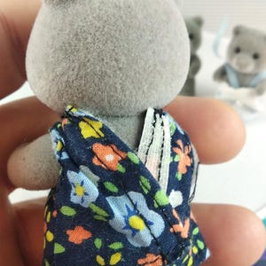 May include: A grey bear figurine wearing a navy blue floral patterned dress. The dress features a colourful floral design with blue, yellow, and orange flowers. Other bear figurines are in the background.