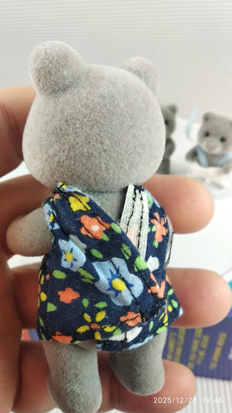 May include: A grey bear figurine wearing a navy blue floral patterned dress. The dress features a colourful floral design with blue, yellow, and orange flowers. Other bear figurines are in the background.