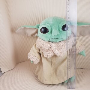 May include: Green and brown plush toy of a character from the Star Wars franchise, The Mandalorian. The toy is wearing a brown hooded robe and has large, dark eyes. The toy is approximately 28 inches tall.