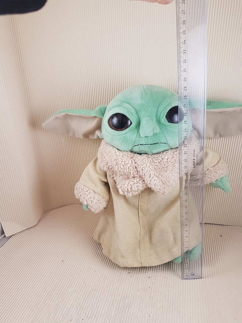 May include: Green and brown plush toy of a character from the Star Wars franchise, The Mandalorian. The toy is wearing a brown hooded robe and has large, dark eyes. The toy is approximately 28 inches tall.