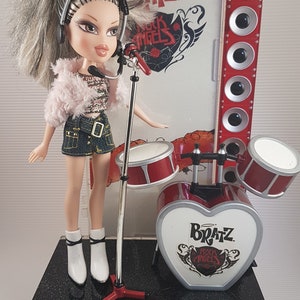Può includere: Una bambola Bratz Rock Angel con un microfono e una batteria su un palco nero. La bambola indossa una giacca rosa, una gonna di jeans e stivali bianchi. La batteria è rossa e bianca e ha una base a forma di cuore con il logo Bratz Rock Angel.