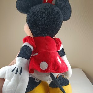 May include: A plush toy of Minnie Mouse, wearing a red dress with white polka dots and a black bow. She has black fur, white gloves, and yellow shoes. Her tail is black and curly.