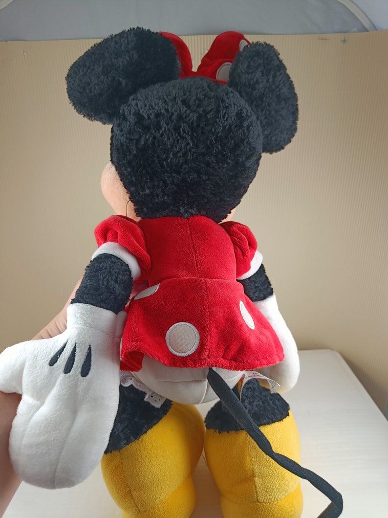 May include: A plush toy of Minnie Mouse, wearing a red dress with white polka dots and a black bow. She has black fur, white gloves, and yellow shoes. Her tail is black and curly.