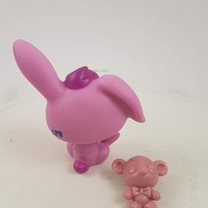 May include: Two small, pink plastic figurines. One is a bunny with large ears and a small purple accent on its head. The other is a small bear with a bow tie. The bunny is approximately 2 inches tall.