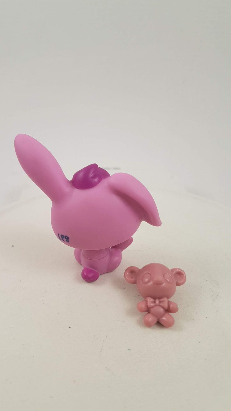 May include: Two small, pink plastic figurines. One is a bunny with large ears and a small purple accent on its head. The other is a small bear with a bow tie. The bunny is approximately 2 inches tall.