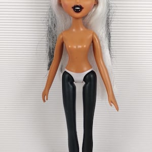 Op de afbeelding: Een Bratz-pop met wit en zwart haar, groene ogen en donkere lippenstift. De pop draagt een witte onderbroek en zwarte leggings. De huidskleur van de pop is bruin.