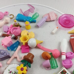 Puede incluir: Una colecci&oacute;n de accesorios de mu&ntilde;ecas en miniatura en varios colores, incluyendo rosa, azul, amarillo y verde. Los art&iacute;culos incluyen biberones, tazas, un plato con detalles florales, un sombrero y un libro titulado "Painting & Drawing".