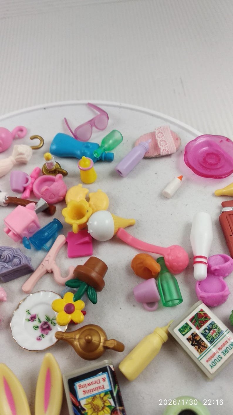 Puede incluir: Una colecci&oacute;n de accesorios de mu&ntilde;ecas en miniatura en varios colores, incluyendo rosa, azul, amarillo y verde. Los art&iacute;culos incluyen biberones, tazas, un plato con detalles florales, un sombrero y un libro titulado "Painting & Drawing".