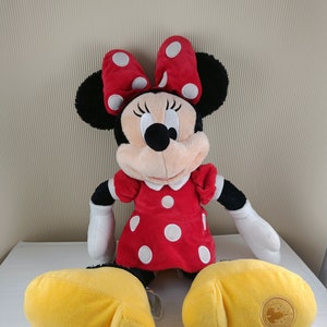 May include: A plush Minnie Mouse toy wearing a red dress with white polka dots and a red bow with white polka dots. The toy has yellow shoes and a Disney Parks logo on one foot.