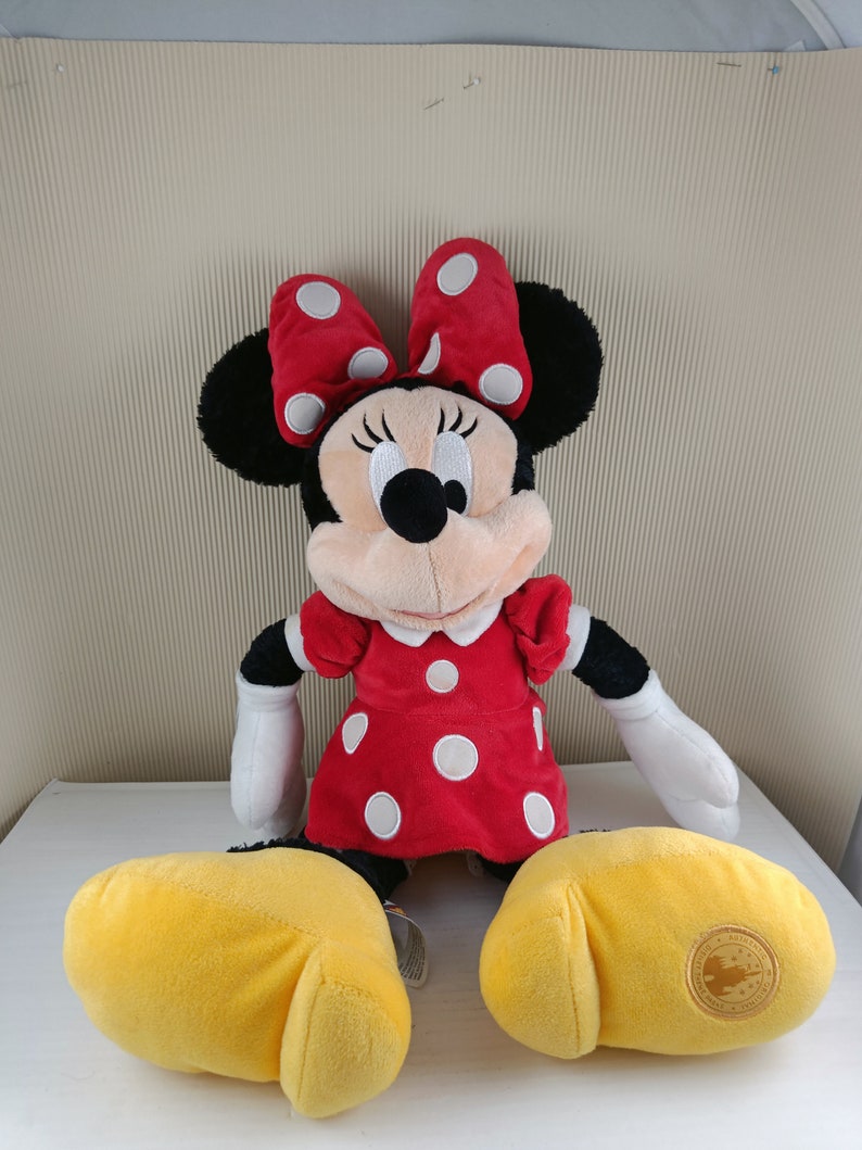 May include: A plush Minnie Mouse toy wearing a red dress with white polka dots and a red bow with white polka dots. The toy has yellow shoes and a Disney Parks logo on one foot.