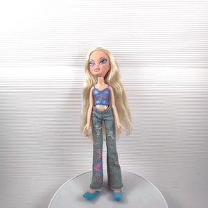 May include: A Bratz doll with long blonde hair, wearing a blue and pink sequined crop top, denim jeans with pink and blue designs, and blue shoes. The doll is standing on a white surface.