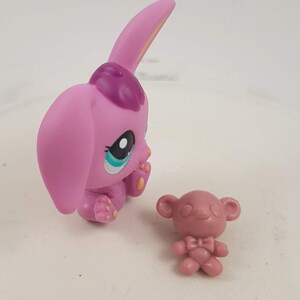 May include: A pink toy bunny figure with large ears and a purple patch on its head, next to a small pink bear figure. The bunny has large eyes with blue and white accents. The figures are on a white surface.