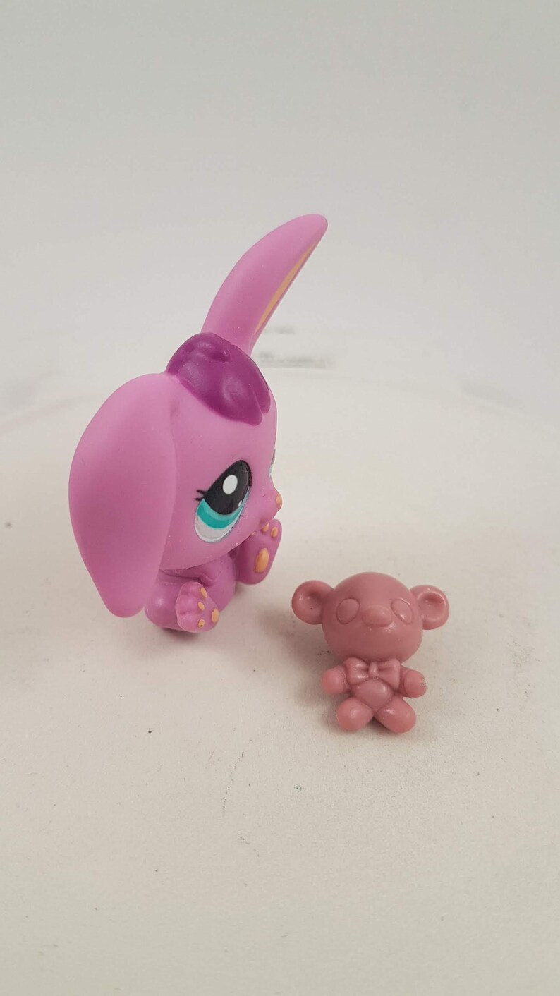 May include: A pink toy bunny figure with large ears and a purple patch on its head, next to a small pink bear figure. The bunny has large eyes with blue and white accents. The figures are on a white surface.