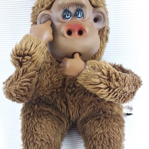 May include: A vintage brown plush monkey toy with a plastic face, hands, and feet. The monkey has a brown fur body, a tan face, and blue eyes. The monkey is in a sitting position with its hands near its face.