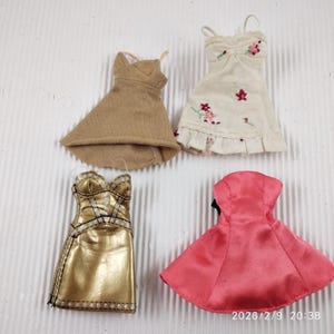 May include: Four miniature dresses in various styles and colors. Includes a tan wrap dress, a white dress with floral embroidery, a gold metallic dress with silver accents, and a pink satin dress.