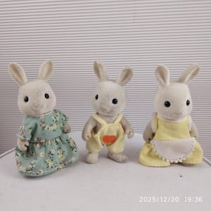 May include: Three small, plush rabbit figurines. One wears a floral print dress, another wears yellow overalls with a carrot, and the third wears a yellow dress with a white apron. The rabbits have black eyes and long ears.