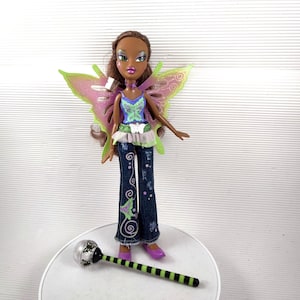 Könnte beinhalten: Eine Modepuppe mit braunen Haaren, grünen und rosa Schmetterlingsflügeln und einem passenden Zauberstab. Die Puppe trägt ein blaues und grünes Oberteil, Jeans mit Schmetterlingsmotiven und lila Schuhe. Der Zauberstab hat einen schwarz-grün gestreiften Griff.