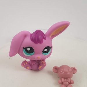 May include: A pink bunny figurine with a dark purple tuft of hair and a small pink bear figurine. The bunny has large eyes with teal accents and yellow feet. The bear has a bow tie.
