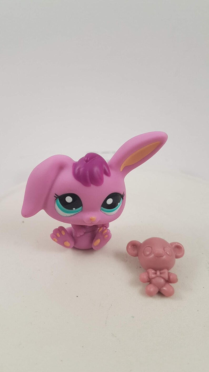 May include: A pink bunny figurine with a dark purple tuft of hair and a small pink bear figurine. The bunny has large eyes with teal accents and yellow feet. The bear has a bow tie.