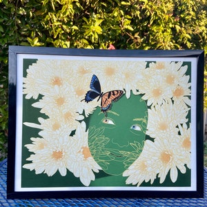 Puede incluir: Una impresión enmarcada de una cara verde rodeada de flores blancas con una mariposa en la frente. La mariposa tiene alas azules y marcas naranjas y negras.