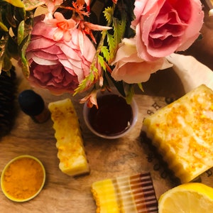 May include: A close-up of a wooden surface with four bars of soap, a small bowl of turmeric powder, a lemon wedge, and a small dish of honey. The soap bars are layered with different colors, including yellow, orange, and brown. The soap bars are decorated with small pieces of dried flowers.