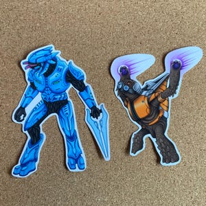 Puede incluir: Dos pegatinas coloridas que representan a un guerrero alienígena azul y negro sosteniendo una cuchilla de energía azul y una criatura parecida a una tortuga marrón y naranja con explosiones de energía púrpura que salen de sus manos.
