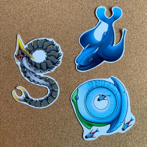 May include: Three colourful stickers featuring illustrations of sea creatures. The first sticker is a black and grey worm-like creature with a yellow and red mouth. The second sticker is a blue fish with a white belly and a black eye. The third sticker is a blue and green fish with a white eye and a circle in the centre.