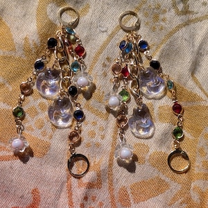 May include: A pair of gold earrings with multiple strands of colourful beads and clear, teardrop-shaped charms. The earrings feature a variety of jewel-toned beads in red, blue, green, and clear, with small white flower-shaped accents.