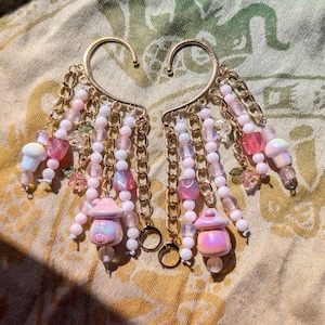 May include: A pair of gold-colored ear cuffs with dangling chains and beads. The chains are adorned with pink, white, and clear beads, along with small mushroom and house charms. The ear cuffs are set against a patterned fabric.