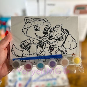 May include: A paint-by-numbers kit featuring two cartoon puppies. The canvas shows the outlines of the characters, with a set of small paint pots in various colors and two paintbrushes. The kit is held in a person's hand.