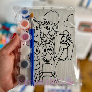 May include: Paint-by-number kit featuring a cartoon dog family. The kit includes a canvas with a black outline drawing, small paint pots with various colors, and two paintbrushes. The canvas is packaged in a clear plastic bag.