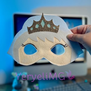 May include: A white felt mask with a gold glitter crown and blue eyes. The mask is designed to look like a princess.