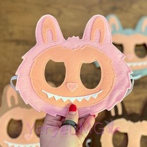 May include: Pink felt monster mask with peach-colored facial features, including a mouth with white teeth, and large ears. The mask has eye holes and an elastic band. Other masks are visible in the background.