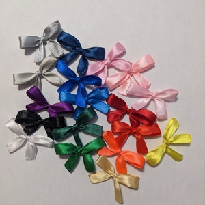 Op de afbeelding: Een verzameling van 18 kleine satijnen strikjes in verschillende kleuren, waaronder wit, zwart, rood, oranje, geel, groen, blauw, paars en roze. De strikjes zijn in een halve cirkel op een witte achtergrond gerangschikt.