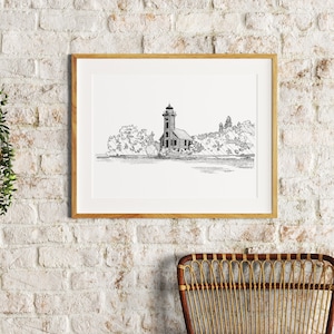 May include: A black and white line drawing of a lighthouse on a small island with trees. The drawing is framed and hanging on a white brick wall. A wicker chair and a woven basket with a brown pillow are in the foreground.