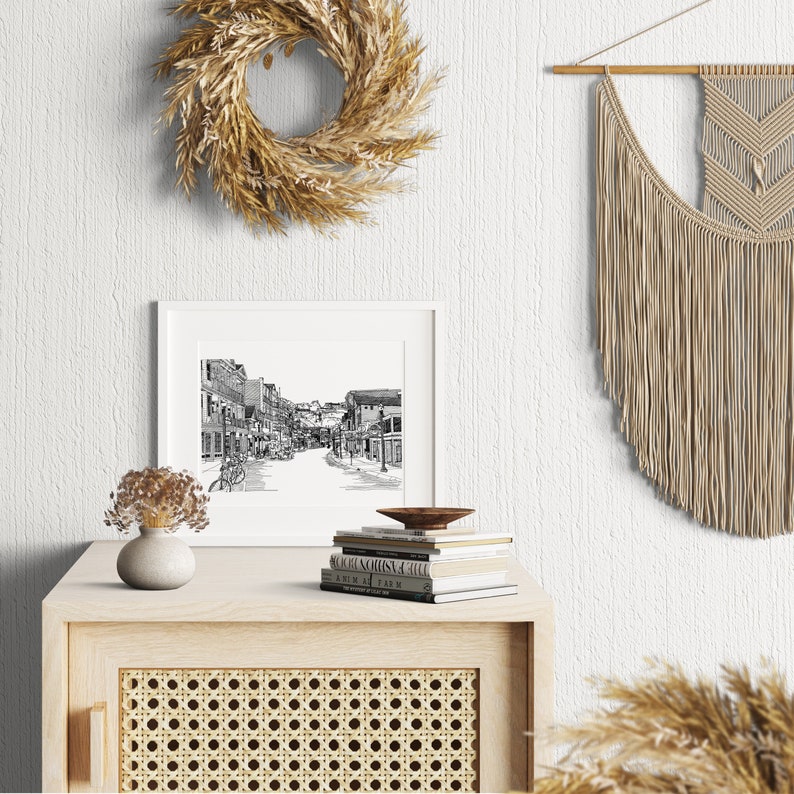 May include: A black and white framed print of a street scene sits on a light wood cabinet with a woven cane door. A stack of books and a small wooden bowl are on top. A dried floral arrangement in a vase and a decorative wreath are also present.