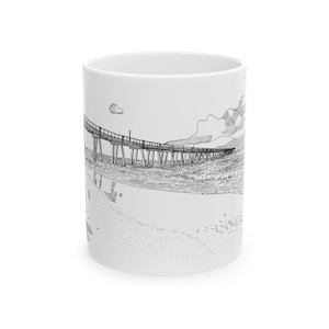 May include: A white ceramic mug featuring a black and white line drawing of a pier over a beach. The pier has a wooden deck and supports, and the beach has sand and waves.