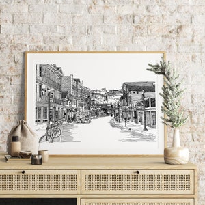 May include: A framed black and white pen and ink drawing of a street scene. The artwork shows a detailed view of a street with buildings, shops, and bicycles. The artwork is displayed on a wooden cabinet, with a vase and decorative items.