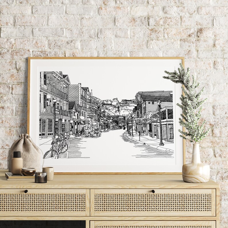 May include: A framed black and white pen and ink drawing of a street scene. The artwork shows a detailed view of a street with buildings, shops, and bicycles. The artwork is displayed on a wooden cabinet, with a vase and decorative items.