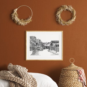 May include: A framed black and white street scene print hangs on a terracotta-coloured wall, with two dried wreath decorations. A woven basket and a beige knitted throw are in the foreground. The print shows a detailed street with buildings and bicycles.