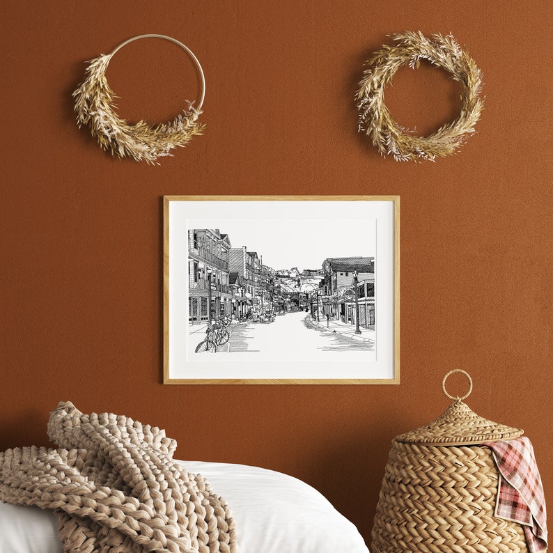 May include: A framed black and white street scene print hangs on a terracotta-coloured wall, with two dried wreath decorations. A woven basket and a beige knitted throw are in the foreground. The print shows a detailed street with buildings and bicycles.