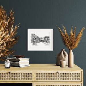 May include: A framed black and white street scene print hangs on a dark teal wall. The print shows a detailed illustration of a street with buildings and people. The print is displayed above a light wood cabinet with decorative items.