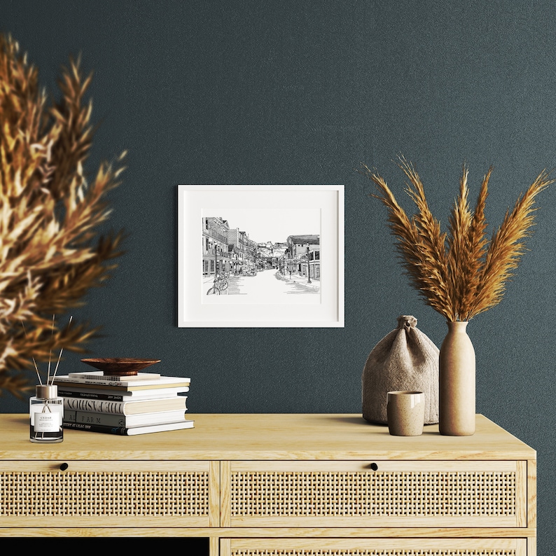 May include: A framed black and white street scene print hangs on a dark teal wall. The print shows a detailed illustration of a street with buildings and people. The print is displayed above a light wood cabinet with decorative items.