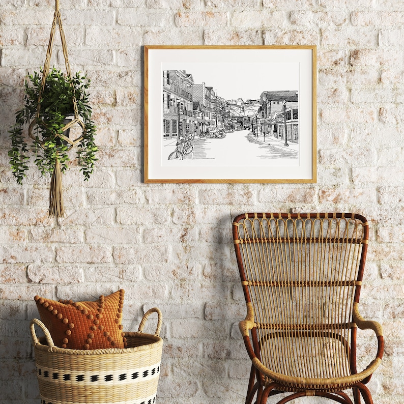 May include: A framed black and white print of a street scene hangs on a white brick wall. The print is in a light wood frame. A woven basket with a decorative cushion and a rattan chair are in front of the wall. A hanging plant is to the left.
