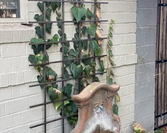 May include: A terracotta fountain with a decorative design and a small pool of water. The fountain is placed in front of a brick wall with a metal trellis covered in green ivy.